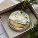 A decorative ornament in a box, featuring a scenic landscape with a beach, trees, and a bird in flight.