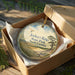 A round ornament in a box, featuring a scenic landscape with a beach, trees, and a bird in flight.