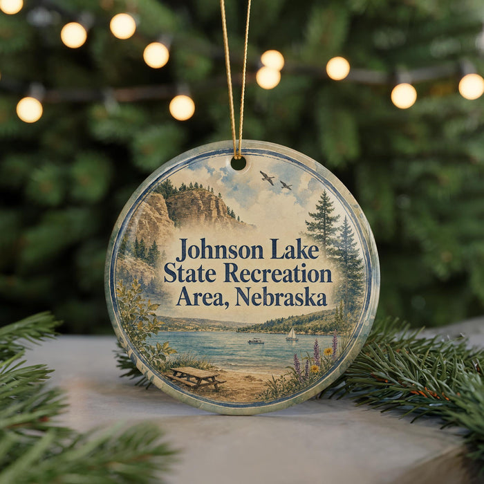 A round ornament with a scenic landscape of Johnson Lake, State Recreation Area, Nebraska, featuring a lake, mountains, pine trees, and a picnic table.