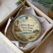 A round ornament with a lighthouse and beach scene, placed in a box with a white tissue paper lining.