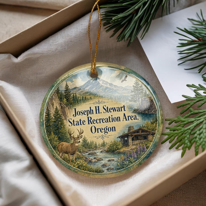 A decorative ornament with a scenic landscape featuring a deer, mountains, and a cabin, hanging from a string.