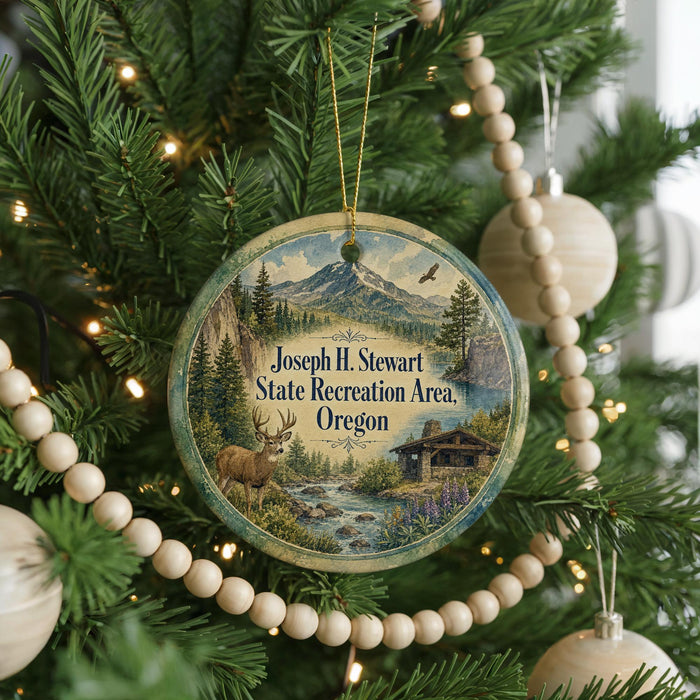 A round ornament hanging on a Christmas tree, featuring a scenic landscape with mountains, a river, and a cabin.