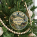 A round ornament hanging on a Christmas tree, featuring a scenic landscape with mountains, a river, and a cabin.