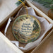 A decorative ornament in a box, featuring a scenic landscape with pine trees, a sailboat, and a body of water.