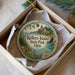 A round ornament with a lighthouse and a scenic landscape, placed in a box with a white tissue paper lining.