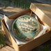 A decorative ornament in a box, featuring a scenic landscape with a lighthouse and a body of water.