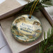 A decorative ornament in a box, featuring a scenic landscape with mountains, pine trees, and a lake.