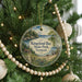 A round ornament hanging on a Christmas tree, featuring a scenic landscape with a house and trees.