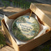A round ornament in a box, featuring a scenic landscape with a house and trees, is displayed on a wooden surface.