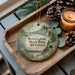 A decorative ornament featuring a scenic landscape of Kinckinnic State Park, Wisconsin, with pine cones and a lit candle on a wooden tray.