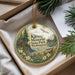 A decorative ornament with a scenic landscape, featuring a cabin, trees, and a lake, is displayed in a box with pine branches.