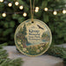 A round ornament with a scenic landscape featuring a cabin, trees, and a bird in flight, hanging from a string.