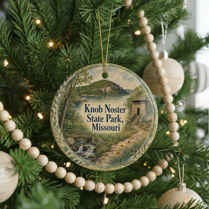 A round ornament hanging on a Christmas tree, featuring a scenic landscape with a bridge and a lake.