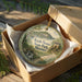 A decorative ornament in a box, featuring a scenic landscape with a bridge and trees, with the text "Knob Noster State Park, Missouri" printed on it.