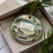 A decorative ornament with a scenic landscape of Kupachuck State Park, Washington, featuring pine trees, a river, and a bird in flight.