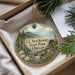 A round ornament with a scenic landscape of mountains, forests, and a small cabin, hanging from a string.