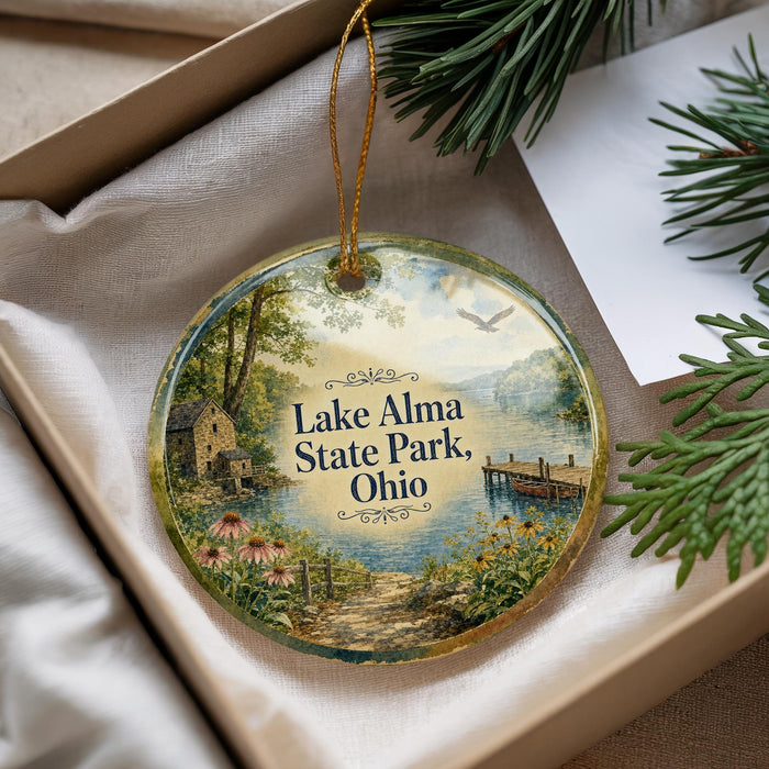 A decorative ornament with a scenic landscape of Lake Alma State Park, Ohio, featuring a small cabin, a lake, and a bird in flight.