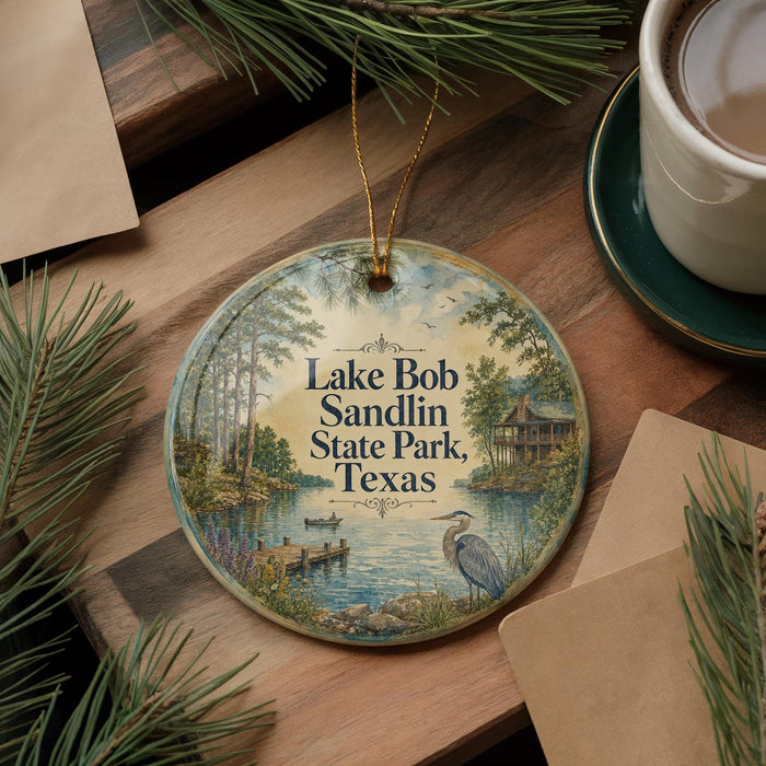 A round ornament with a painting of Lake Bob Sandlin State Park in Texas, featuring a bird and a dock, is displayed on a wooden surface alongside a cup of coffee and some pine branches.