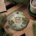 A round ornament with a painting of Lake Bob Sandlin State Park in Texas, featuring a bird and a dock, is displayed on a wooden surface alongside a cup of coffee and some pine branches.