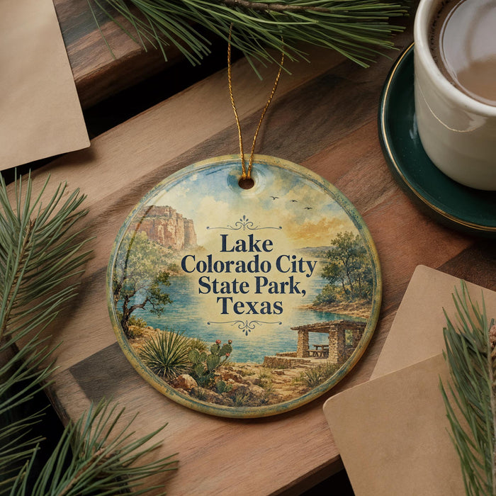 A round ornament with a scenic landscape of Lake, Colorado City, State Park, and Texas, featuring a desert and a lake, is displayed on a wooden surface alongside a cup of coffee and some pine branches.
