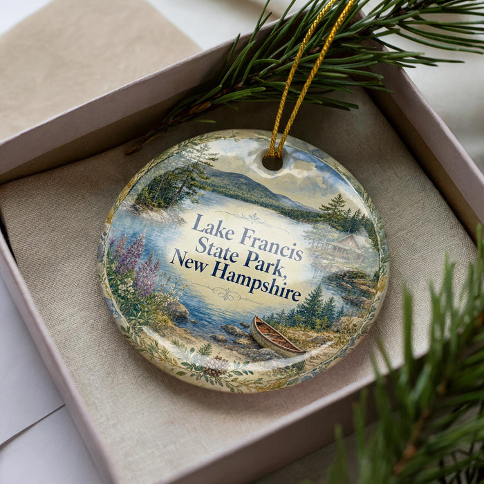 A decorative ornament in a box, featuring a scenic landscape with a lake, mountains, and pine trees.