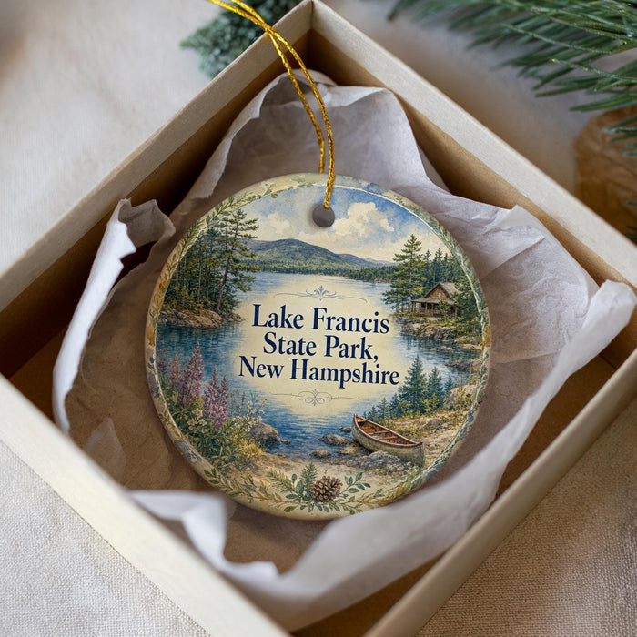 A decorative ornament in a box, featuring a scenic landscape with a lake, mountains, and a cabin.