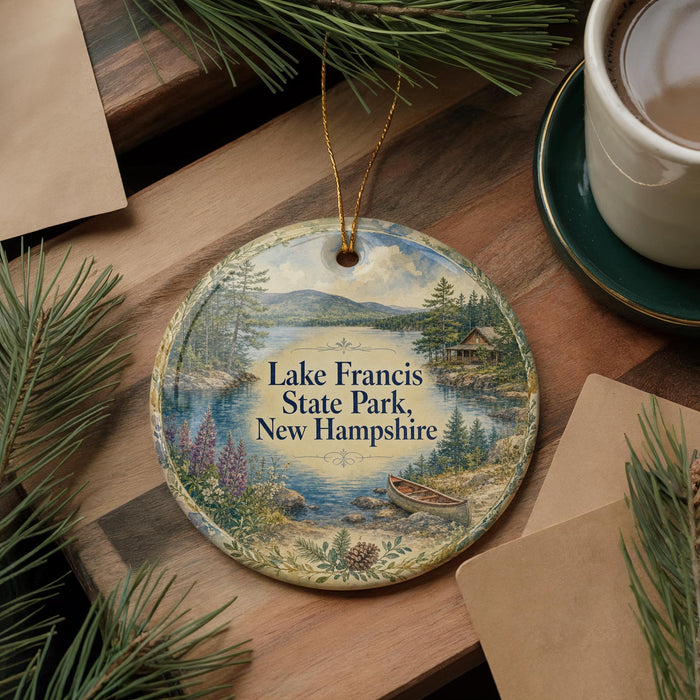 A decorative ornament featuring Lake Francis State Park in New Hampshire, with a scenic landscape and a small boat on the water. The ornament is hanging on a wooden surface, surrounded by pine branches and a cup of coffee.
