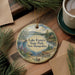 A decorative ornament featuring Lake Francis State Park in New Hampshire, with a scenic landscape and a small boat on the water. The ornament is hanging on a wooden surface, surrounded by pine branches and a cup of coffee.