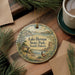A decorative ornament with a scenic landscape of Lake Herman State Park, South Dakota, featuring a bird and a cabin, is displayed on a wooden surface alongside a cup of coffee and some pine branches.