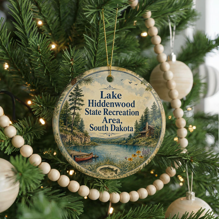 A round ornament hanging on a Christmas tree, featuring a scenic landscape with a lake, trees, and a cabin.