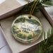 A decorative ornament in a box, featuring a scenic landscape with a lake, trees, and wildlife, including a bird and a deer.