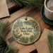 A round ornament with a scenic landscape of Lake Logan State Park, Ohio, featuring a bird, deer, and a cabin, is displayed on a wooden surface alongside a cup of coffee and some pine branches.