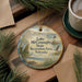 A round ornament with a scenic lake and lighthouse image, surrounded by pine branches and a cup of coffee on a wooden surface.