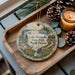 A decorative ornament featuring a scenic landscape of Lake Metigoshe State Park in North Dakota, with pine cones and a lit candle on a wooden tray.