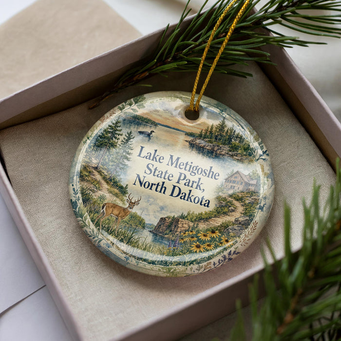 A decorative ornament featuring a scenic landscape of Lake Metigoshe State Park, North Dakota, with a deer and pine trees in the foreground.