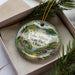 A decorative ornament in a box, featuring a scenic landscape with a lake, trees, and a waterfall.