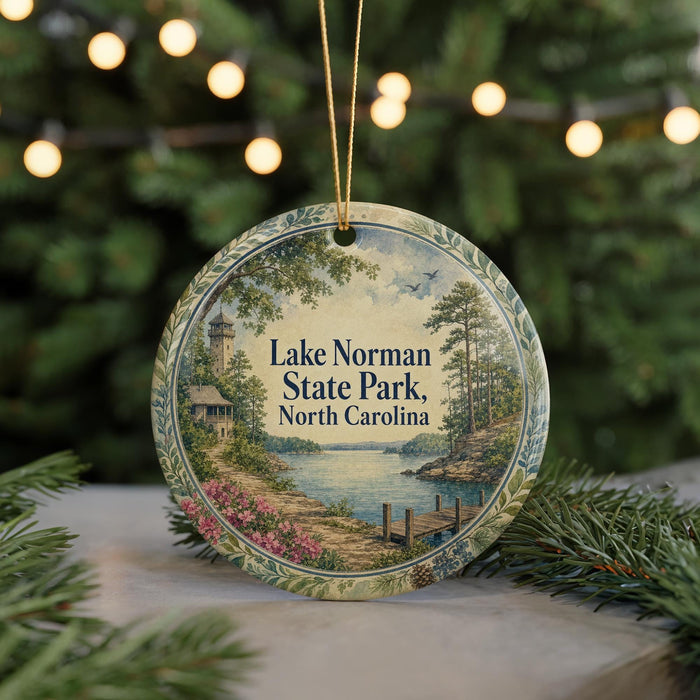 A round ornament with a scenic landscape of Lake Norman State Park, North Carolina, featuring a lighthouse, trees, and a body of water.
