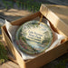 A decorative ornament in a box, featuring a scenic landscape with a lake, trees, and a bridge.