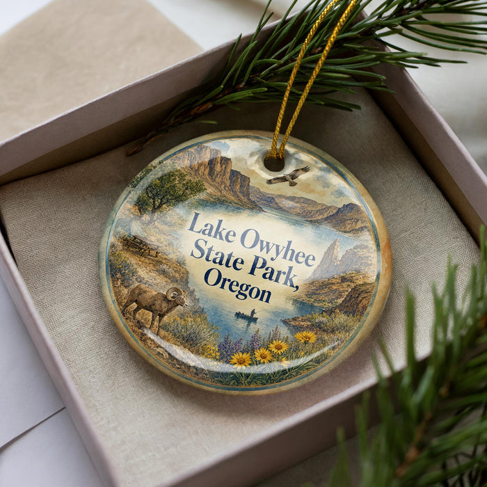 A decorative ornament in a box, featuring a scenic landscape with mountains, a river, and various wildlife elements, including a bird and a deer.