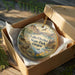 A decorative ornament in a wooden box, featuring a landscape scene with mountains, a river, and various wildlife elements.