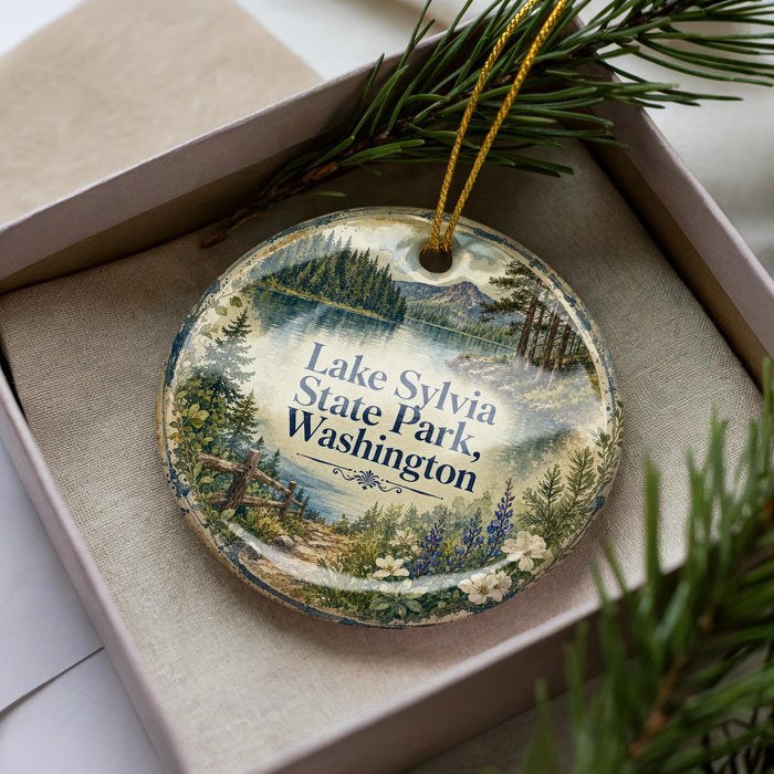 A decorative ornament in a box, featuring a scenic landscape with pine trees, a lake, and mountains.