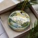 A decorative ornament with a scenic lake and forest landscape, featuring a boat on the water, is displayed in a box with pine branches.