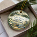 A decorative ornament with a lake and park scene, featuring a bird and a boat, is displayed in a box with pine branches.