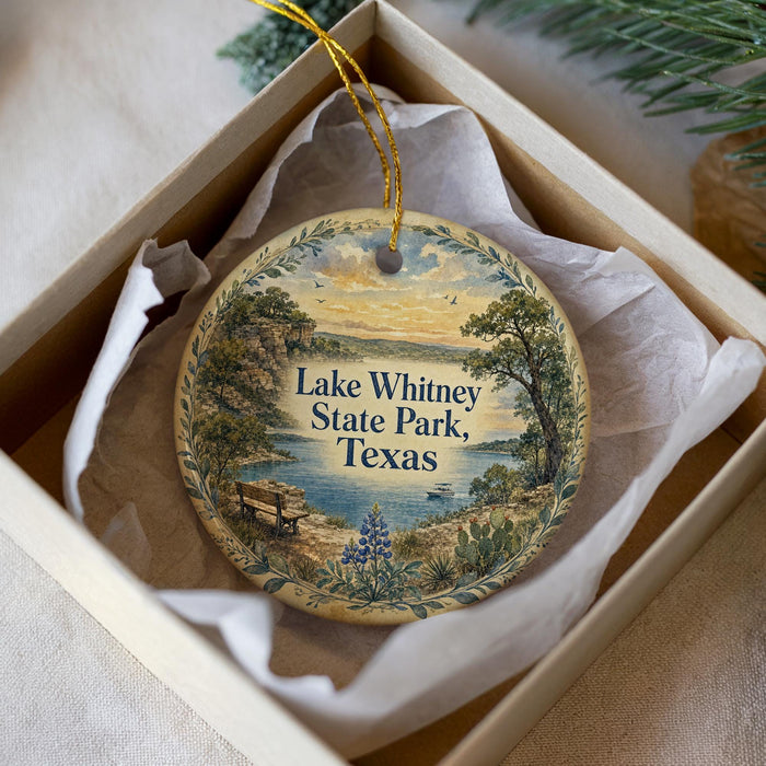 A decorative ornament in a white box, featuring a scenic landscape with a lake, trees, and a bench.