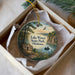 A decorative ornament in a box, featuring a scenic landscape with a lake, a cabin, and a bird in flight.