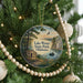 A round ornament hanging on a Christmas tree, featuring a scenic landscape with a lake, trees, and a cabin.