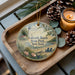 A decorative ornament featuring a lighthouse and a beach scene, placed on a wooden tray alongside pine cones and a lit candle.