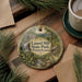 A round ornament with a scenic image of Laurel Hill State Park in Pennsylvania, featuring a waterfall, cabin, and forest. The ornament is hanging from a string and is placed on a wooden surface, surrounded by pine branches and a cup of coffee.