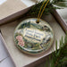 A decorative ornament in a box, featuring a scenic landscape with pine trees, a waterfall, and a bird.