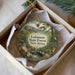 A round ornament with a scenic forest landscape and a bird perched on a branch, placed in a box with a white tissue paper lining.
